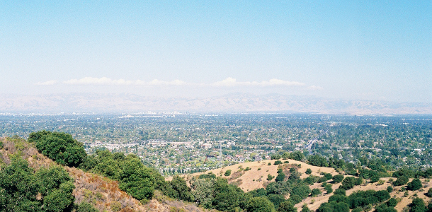 [Two images stitched together showing more of the hilltops in the foreground and more of the valley below and the mountains in the distance. There is a string of white clouds which appear to be over the distant mountains. No haze over the valley, so the streets and trees in the valley are more distinct.]